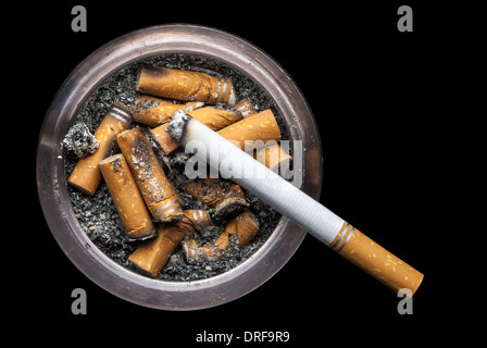 Eine Grunge Aschenbecher voller Asche und Zigarettenkippen mit einer brennenden Zigarette auf einem schwarzen Hintergrund und Platz für Kopie. Stockfoto