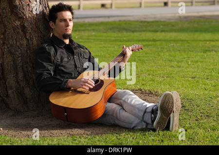 Ein weißer junge Mann sitzen, lehnte sich gegen einen Baum, die Gitarre in einem park Stockfoto