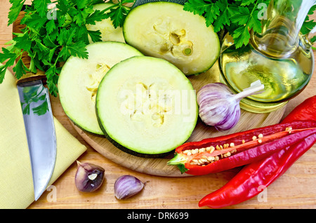 Zucchini, Knoblauch, Pflanzenöl in Karaffe, Serviette, Messer auf einem Holzbrett, heiße rote Paprika, Petersilie Stockfoto