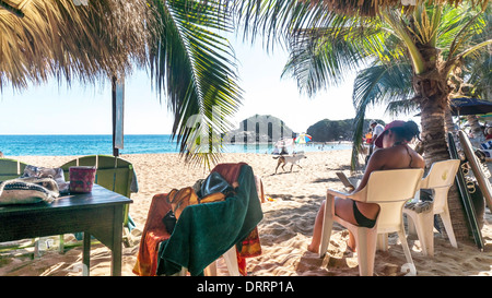 Frau genießt das gute Leben lesen unter Palapa Dach der beliebte Strandrestaurant mit Blick auf Menschen, die Schwimmen im Meer Stockfoto