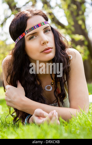 Hübsche junge Frau mit schöne lange gewellte brünette Haare in einem Stirnband liegen auf den frischen grünen Sommer Rasen träumen in der Sonne mit einem fernen Ausdruck Stockfoto