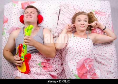 Junges Paar im Bett liegend. Mann spielt Gitarre und Schreie seiner Stimme. Frau bedeckt ihre Finger in die Ohren Stockfoto