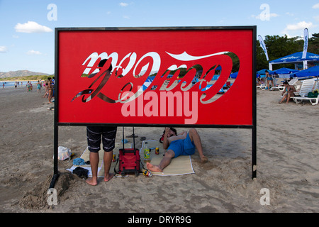 Junge Menschen stehen und sitzen als nächstes ein Coca Cola Schild am Strand von Playa Tamarindo, Guanacaste, Costa Rica Stockfoto