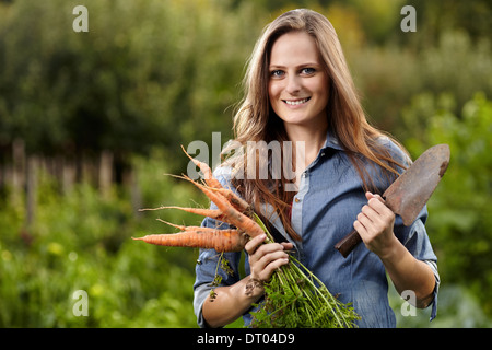 Junge Frau Gärtner hält eine Garbe von Möhren und eine Hacke in ihrem Garten, selektiven Fokus Stockfoto