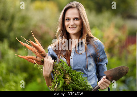 Junge Frau Gärtner hält eine Garbe von Möhren und eine Hacke in ihrem Garten, selektiven Fokus Stockfoto
