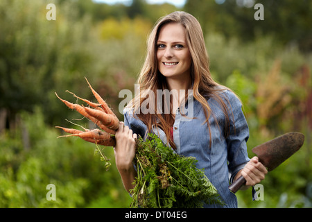 Junge Frau Gärtner hält eine Garbe von Möhren und eine Hacke in ihrem Garten, selektiven Fokus Stockfoto