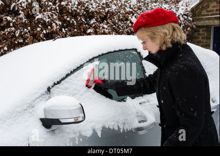 HERTFORD, UK - 18. Januar 2013: Eine Frau in einem roten Hut löscht Schnee aus den Fenstern eines Silber farbigen Autos bei starkem Schneefall. Stockfoto