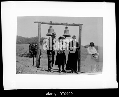 Dieses Foto zeigt die Taufe eines Babys im Freien, die um 1910 unter den alten Missionsglocken stattfindet und einen Moment der religiösen Zeremonie in einer historischen Umgebung festhält. Stockfoto