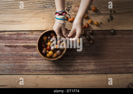 Eine Runde Schale mit kleinen Tomaten, verschiedene Farben und eine Person, Sortierung und Kommissionierung über sie eine hölzerne Tischplatte Stockfoto