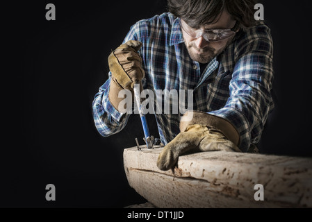 Ein Mann arbeitet in einem aufgearbeiteten Holzplatz Workshop Besitz Werkzeuge und Nägel aus einem verknotet und unebene Stück Holz Stockfoto