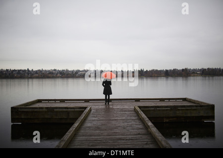 Steht eine Frau trägt alle schwarz und orangenen Regenschirm hält am Rande eines Docks auf eine graue und trübe Tag in Seattle WA Stockfoto