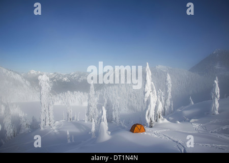 Ein helles orange Zelt unter Schnee bedeckt Bäume auf einem verschneiten Bergrücken mit Blick auf einen Berg in der Ferne Stockfoto