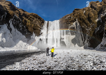 Regenbogen über Skogafoss Wasserfall im Winter Island Paar genießt die Nähe zu den Wasserfall mit einem schönen Regenbogen. Stockfoto