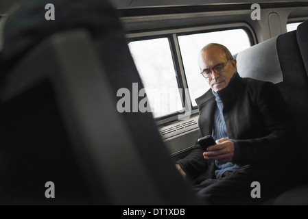 Ein reifer Mann sitzt an einem Fenster in einem Zug Wagen mit seinem Mobiltelefon in Kontakt in Bewegung zu bleiben Stockfoto