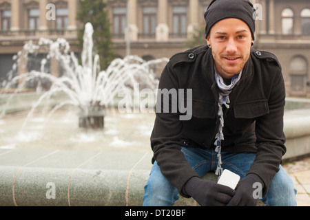Junger lächelnder Mann mit Handy vor Fountan, Stadtraum Stockfoto