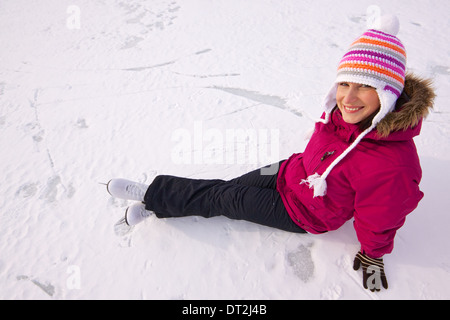 Lächelnde junge Frau mit dem Schlittschuhe tragen gestrickte Wintermütze sitzen im Schnee auf zugefrorenen See Stockfoto