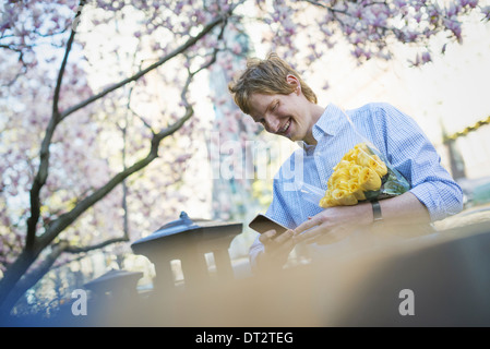 Ein junger Mann im Park im Frühjahr mit einem Mobiltelefon hält eine Reihe von gelben Rosen Stockfoto