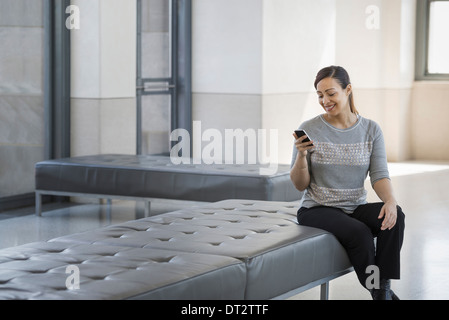 Urban Lifestyle A junge Frau sitzt auf einem Sitzplatz in einem Gebäude mit ihrem Mobiltelefon Stockfoto