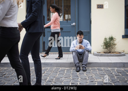 Junge Menschen im Freien auf den Straßen der Stadt im Frühling A Mann sitzt auf dem Boden Überprüfung sein Telefon und drei Passanten Stockfoto