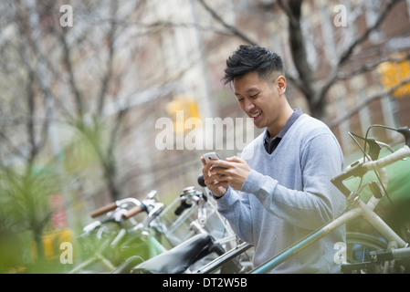 Blick über CityA Mann in einem blauen Pullover mit einer Reihe von abgestellten Fahrrädern überprüfen seine Nachrichten auf einem Smartphone Stockfoto