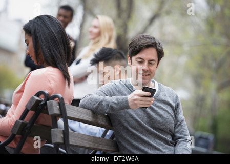 Blick über Citycity Park auf einer Parkbank sitzen, fünf Menschen Männer und Frauen, die ihre Telefone überprüfen Stockfoto