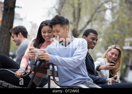 Blick über die Stadt auf einer Bank saß einen Mann zeigt seine Handy-Bildschirm, eine Frau und drei weitere Personen, die ihre Telefone überprüfen Stockfoto