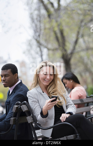 Blick über CityYoung Menschen im Freien in einem Stadt Park drei Leute sitzen auf einer Bank zwei überprüfen ihre Telefone Stockfoto