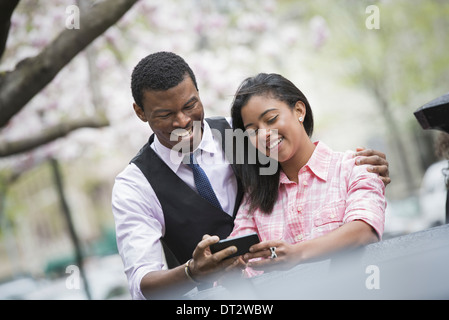Blick über CityYoung Menschen ein paar Seite an Seite mit seinen Arm um ihre Schultern mit Blick auf ein Smartphone und lächelnd Stockfoto