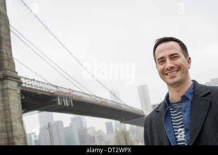 New York City der Brooklyn Bridge überqueren den East River A Mann trägt einen grauen Mantel lächelt in die Kamera Stockfoto