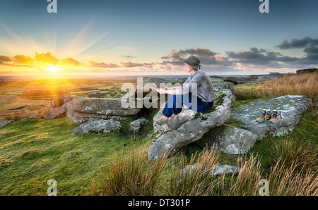 Frau, Wandern, entspannen und unter Verwendung eines Tablet PCs bei Sonnenuntergang auf einem felsigen Tor auf Bodmin Moor in Cornwall Stockfoto