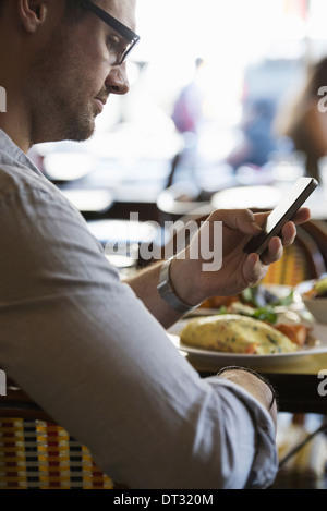 Einen Mann in Freizeitkleidung, eine Café-Tisch Überprüfung seiner Smartphone in Kontakt in Bewegung zu bleiben Stockfoto