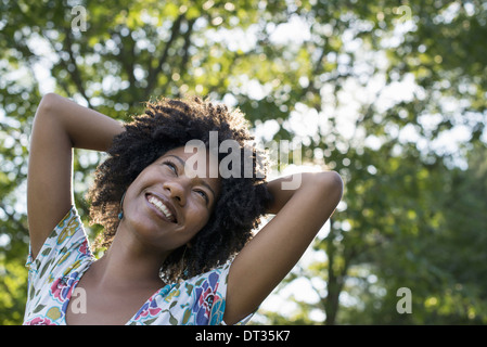 Eine junge Frau in einem geblümten Sommerkleid mit ihren Händen hinter ihrem Kopf lächelnd und blickte Stockfoto