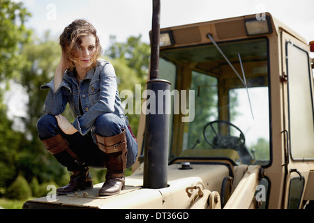 Eine junge Frau in Jeansjacke und Stiefel auf der Motorhaube eines Traktors Stockfoto