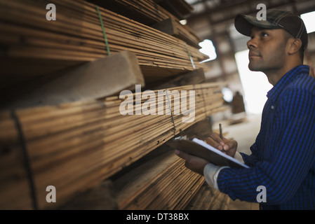 Ein Haufen von recycelten aufgearbeiteten Holzbohlen der Holzplatz. Ein Mann mit einer Zwischenablage durch ein Rack mit Planken. Stockfoto