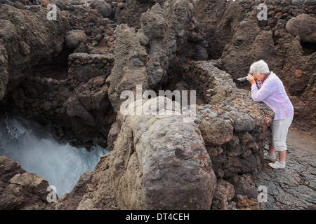 Ältere Frau Tourist fotografiert Wellen in einer Meereshöhle von Weg in Lavagestein Los Hervideros Lanzarote Kanarische Inseln Stockfoto