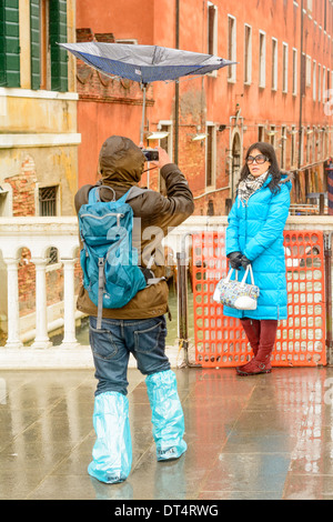 Venedig, Italien. Mann trägt ein umgekehrtes Dach, nimmt Bilder eines asiatischen Frauen in Regenkleidung auf einer Brücke. Stockfoto