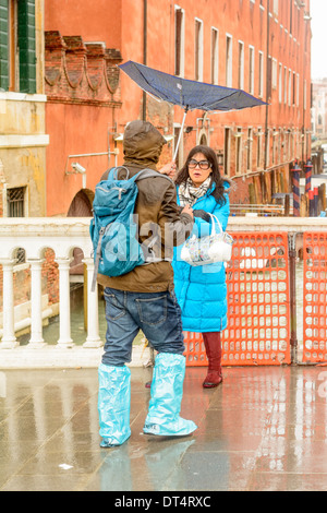 Venedig, Italien. Mann trägt ein umgekehrtes Dach, nimmt Bilder eines asiatischen Frauen in Regenkleidung auf einer Brücke. Stockfoto