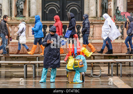 Venedig, Italien. Menschen zu Fuß auf der Acqua Alta Gangway, Verkäufer verkaufen Regenkleidung und Regen Stiefel auf St. Marks Platz. Stockfoto