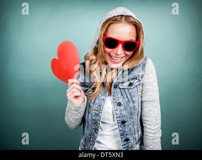 Fröhliches romantische Mädchen Lachen auf blauem Hintergrund, tragen modische Jacke mit Kapuze und Sonnenbrille, roten herzförmigen Karte Stockfoto