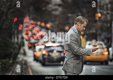 Ein Mann in einem Anzug Überprüfung sein Handy, stehend auf einer belebten Straße in der Abenddämmerung. Stockfoto