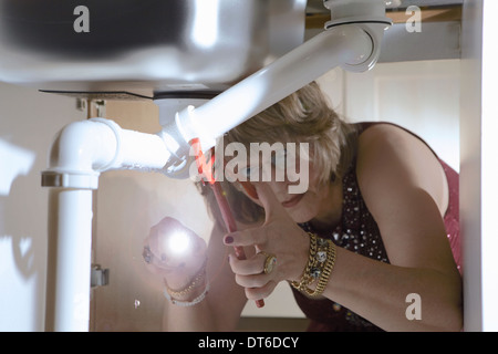 Ältere Frau in Abendgarderobe, die Fixierung der Rohre unter Küchenspüle Stockfoto