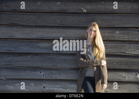 Eine junge Frau lehnt sich an die Holzwand einer Scheune ein langer Strickmantel tragen. Stockfoto