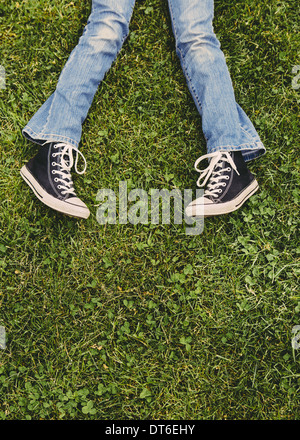 Ein zehn Jahre altes Mädchen auf dem Rasen liegen. Blick auf ihre Unterschenkel beschnitten. Tragen von Turnschuhen und verwaschene blaue Jeans. Stockfoto