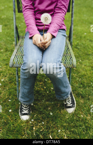 Ein zehnjähriges Mädchen sitzen im Stuhl auf dem grünen Rasen, hält ein Löwenzahn Uhr Seedhead. Verkürzte Ansicht vom Hals abwärts. Stockfoto