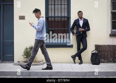 Junge Menschen im Freien auf den Straßen der Stadt im Frühling.  Ein Mann lehnt sich an eine Wand und eine Überprüfung seiner Telefon vorbeigehen. Stockfoto