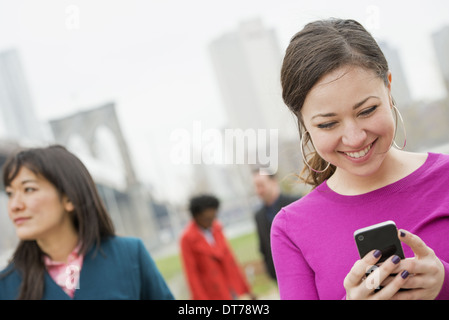 Übergang über den East River. Vier Freunde im Park am Fluss, eine Frau mit Blick auf ihr Handy und lächelnd. Stockfoto
