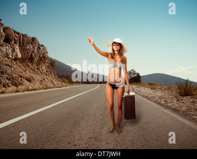 Hübsche junge Frau mit Koffer Trampen entlang einer Straße Stockfoto