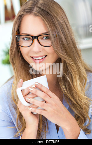 Schöne junge blonde Frau zu Hause in ihrer Küche eine Brille Geek Lächeln und eine Tasse Tee oder Kaffee trinken Stockfoto