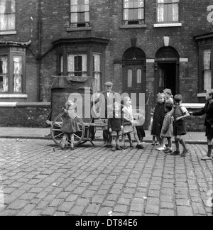 Historisches Bild aus den 1950er Jahren, das eine Gruppe innerstädtischer Kinder in einer gepflasterten Straße zeigt, in der ein älterer Mann mit Knochen und Fetzen neben seinem Handwagen steht. Der Mann war ein Händler, der unerwünschte und weggeworfene Haushaltsgegenstände einsammelte - alte Kleidung, Fahrräder, Metallgegenstände, Möbelstücke usw. - und sie dann weiterverkaufte. Ein ein-Mann-Recycler. Stockfoto