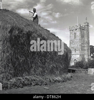 Historisches Bild von 1950 zeigt ein Landwirt mit einer Heugabel auf einem großen Heuhaufen auf einem Feld neben einer Kirche, England. Stockfoto